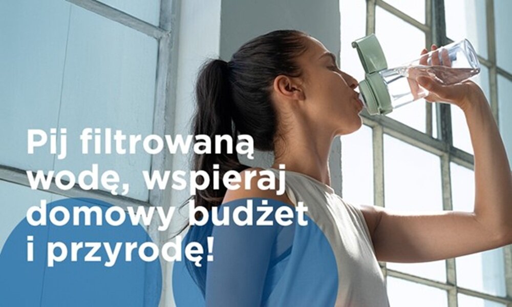 Osoba pije wodę z butelki z filtrem, stojąc przy oknie. Na obrazie widoczny jest tekst: \'Pij filtrowaną wodę, wspieraj domowy budżet i przyrodę!\'.