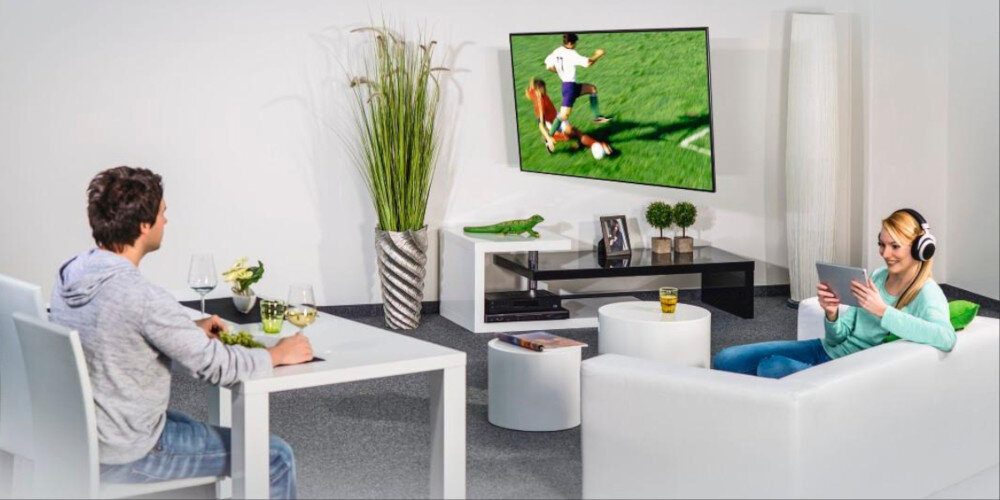 W salonie znajduje się telewizor zamontowany na ścianie, na którym wyświetlany jest mecz piłki nożnej. Dwie osoby siedzą na białych meblach, jedna z nich używa tabletu, a druga je posiłek.