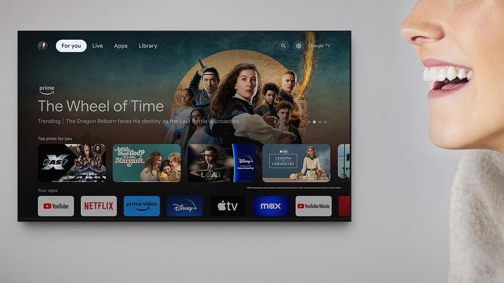 Na ekranie telewizora wyświetlony jest interfejs Google TV z widocznymi aplikacjami takimi jak YouTube, Netflix, Prime Video, Disney+, Apple TV, i max. Na górze ekranu znajduje się grafika promująca serial \'The Wheel of Time\'.