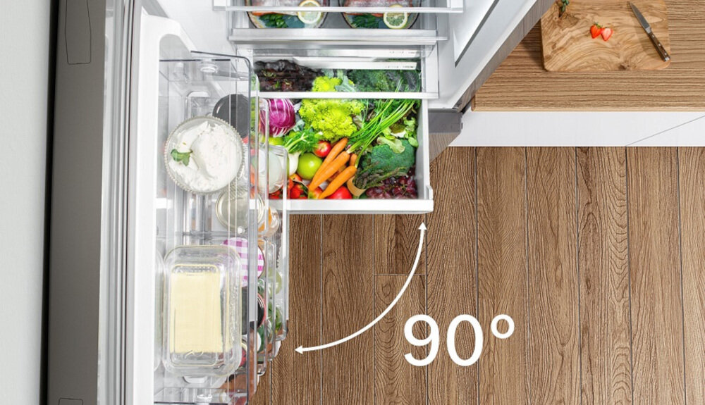 Otwarta lodówka z widocznymi szufladami pełnymi warzyw i produktów spożywczych. Na podłodze obok lodówki znajduje się napis \'90°\'.