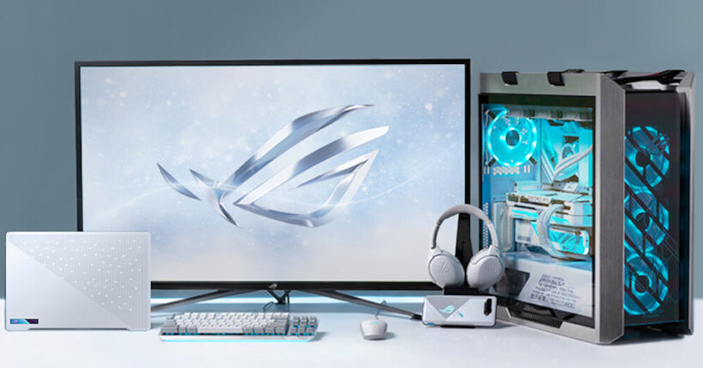 Na obrazie znajduje się zestaw komputerowy składający się z monitora, obudowy komputera z podświetleniem LED, laptopa, klawiatury, myszki, słuchawek i kontrolera. Na ekranie monitora widoczne jest logo ROG.