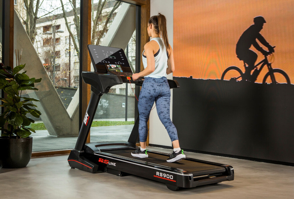 Osoba ćwiczy na bieżni elektrycznej BH FITNESS RS Line RS900 w jasnym pomieszczeniu z dużymi oknami. Na ścianie widoczny jest cień rowerzysty.