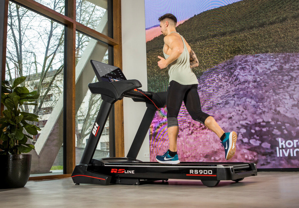 Mężczyzna biegnie na bieżni elektrycznej BH FITNESS RS Line RS900, która znajduje się w jasnym pomieszczeniu z dużymi oknami. Na bieżni widoczne są napisy \'RS Line\' i \'RS900\'.