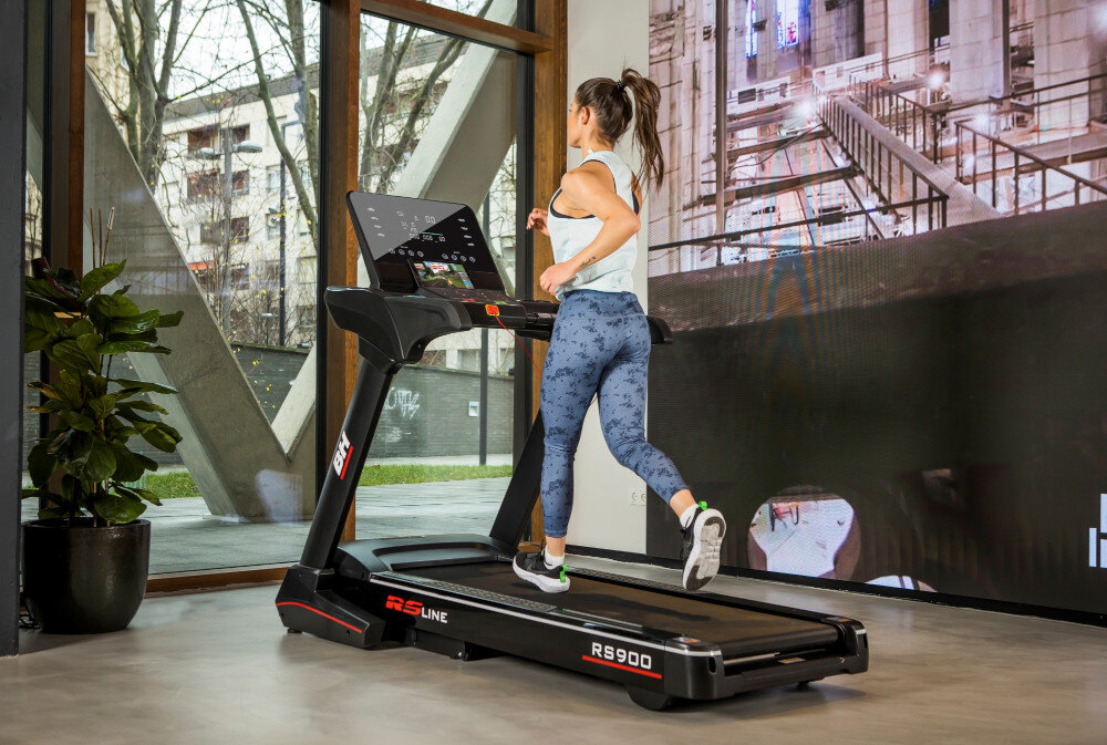 Kobieta biegnie na bieżni elektrycznej BH FITNESS RS Line RS900 w jasnym pomieszczeniu z dużymi oknami. Na bieżni widoczne są napisy \'RS Line\' i \'RS900\'.