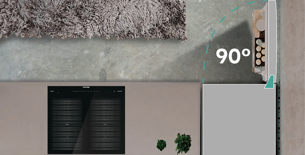 Widok z góry na otwartą lodówkę z napisem \'90°\' oraz fragment dywanu i podłogi. Obok lodówki widoczny jest panel z przyciskami i kilka roślin doniczkowych.