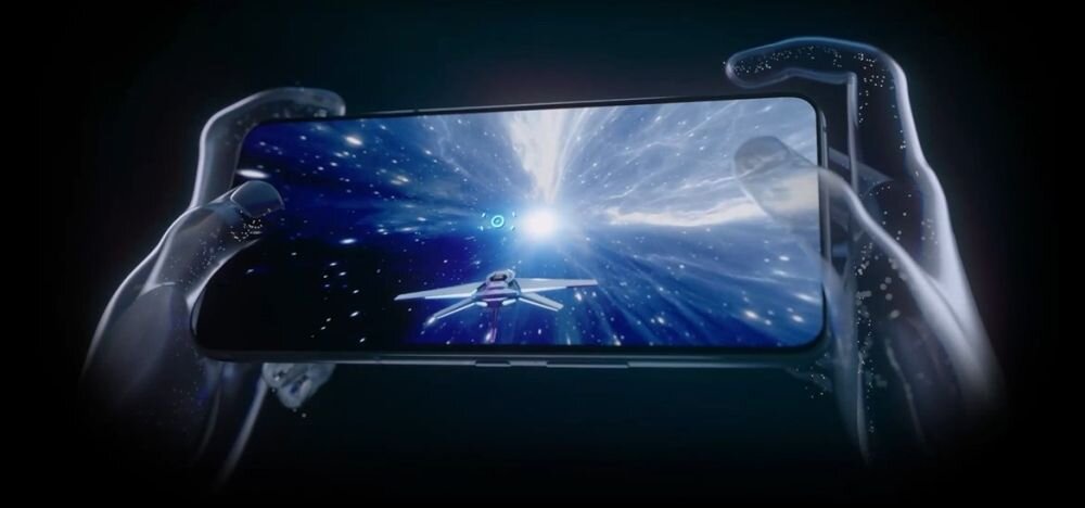Obraz przedstawia smartfon trzymany w przezroczystych dłoniach, na ekranie widoczna jest scena z gry kosmicznej z gwiazdami i statkiem kosmicznym. Tło jest ciemne, co podkreśla jasność ekranu.