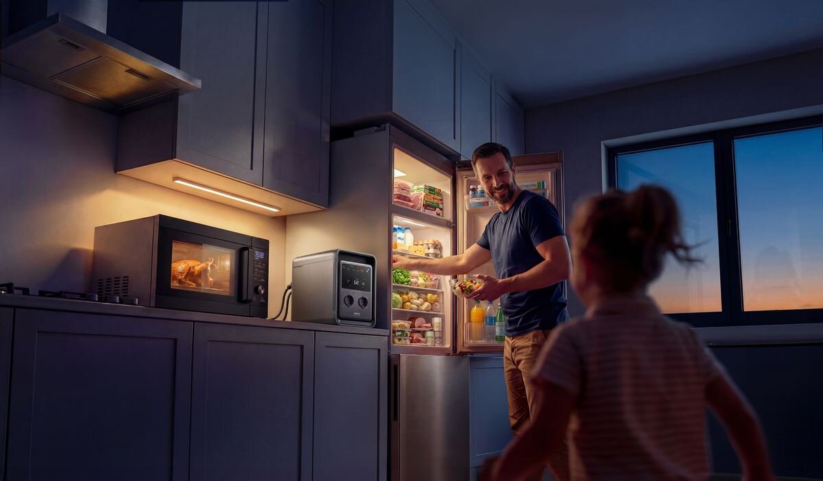 W kuchni znajduje się otwarta lodówka z widocznymi produktami spożywczymi, obok której stoi mężczyzna. Na blacie obok mikrofalówki znajduje się stacja zasilania.