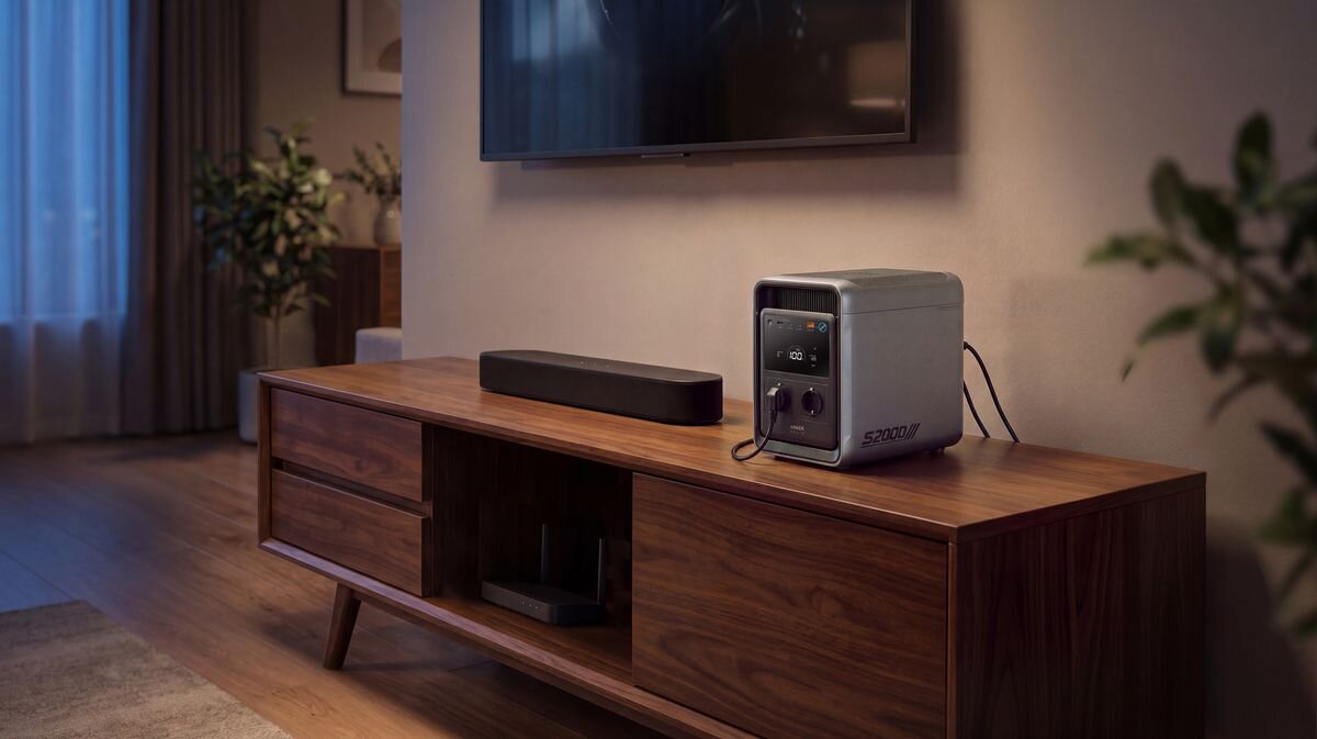 Na drewnianej szafce znajduje się stacja zasilania z wyświetlaczem i dwoma gniazdami, obok której leży czarny soundbar. W tle widać telewizor zawieszony na ścianie oraz rośliny w doniczkach.
