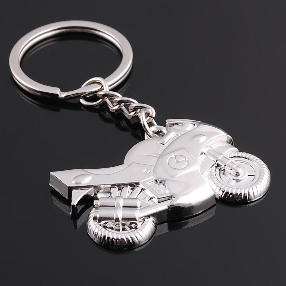 Metalowy brelok do kluczy w kształcie motocykla z detalami opon i kierownicy, przymocowany do kółka na klucze za pomocą łańcuszka. Powierzchnia breloka jest błyszcząca i gładka.