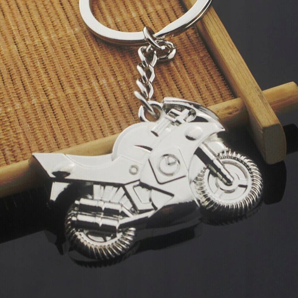 Metalowy brelok do kluczy w kształcie motocykla, leży na drewnianej powierzchni. Brelok jest srebrny i ma szczegółowe wykończenie kół i ramy motocykla.
