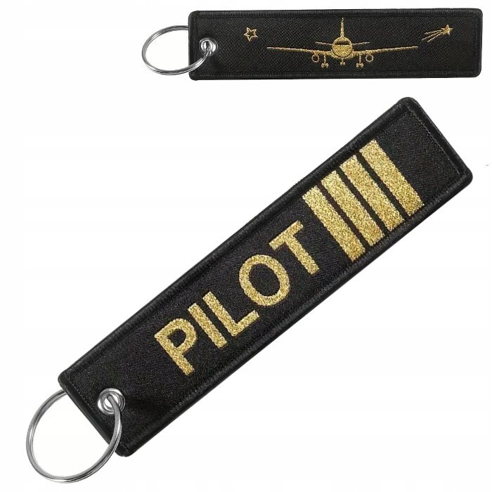 Na obrazie widoczny jest czarny brelok do kluczy z metalowym kółkiem, ozdobiony złotym napisem \'PILOT\' oraz trzema paskami. Na drugiej stronie znajduje się złoty wizerunek samolotu z gwiazdką i strzałką.