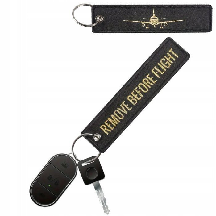 Na obrazie widoczny jest czarny brelok do kluczy z haftem przedstawiającym samolot oraz napisem \'REMOVE BEFORE FLIGHT\'. Brelok jest przymocowany do kluczy i pilota samochodowego.