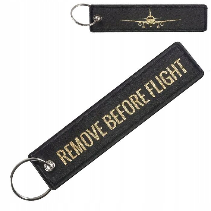 Na obrazie widoczny jest czarny brelok do kluczy z metalowym kółkiem, z napisem \'REMOVE BEFORE FLIGHT\' w kolorze złotym. Na mniejszym breloku znajduje się złoty wizerunek samolotu.
