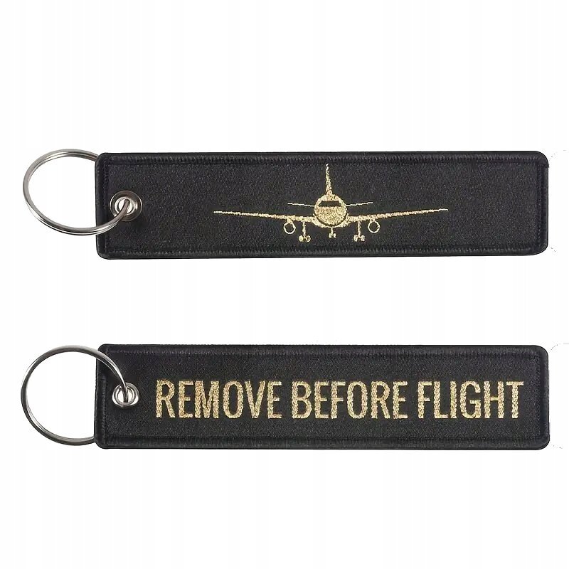 Na obrazie widoczne są dwa czarne breloki do kluczy z metalowymi kółkami. Jeden z nich ma złoty napis \'REMOVE BEFORE FLIGHT\', a drugi złoty wizerunek samolotu.