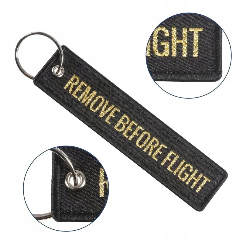 Czarny brelok do kluczy z napisem \'REMOVE BEFORE FLIGHT\' w kolorze złotym, z metalowym kółkiem do mocowania.