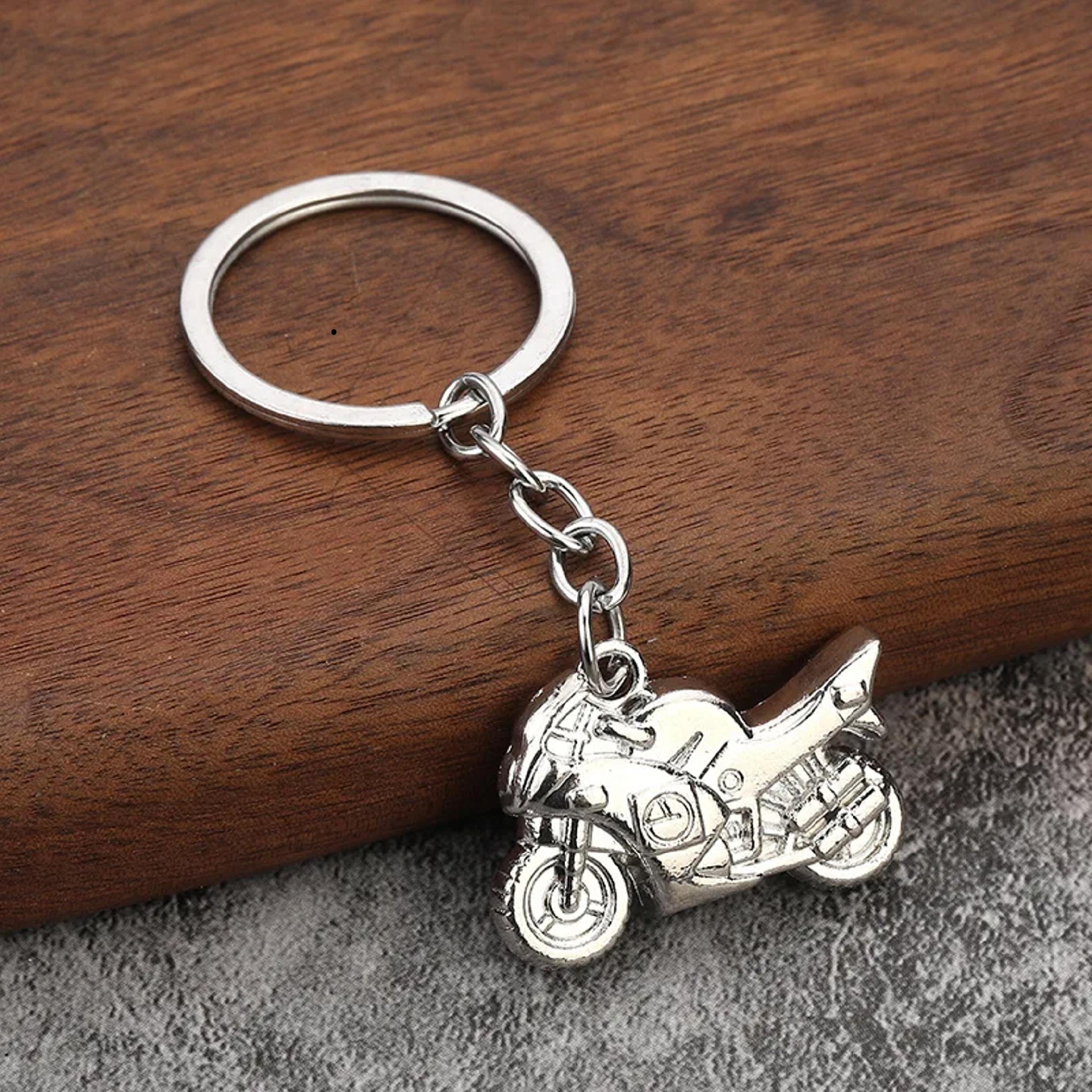 Srebrny metalowy brelok w kształcie motocykla, przymocowany do kółka na klucze za pomocą łańcuszka. Leży na drewnianej powierzchni.