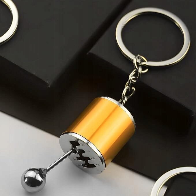 Złoty brelok do kluczy w kształcie skrzyni biegów z metalową kulką na końcu. Brelok jest przymocowany do metalowego kółka na tle czarnych pudełek.