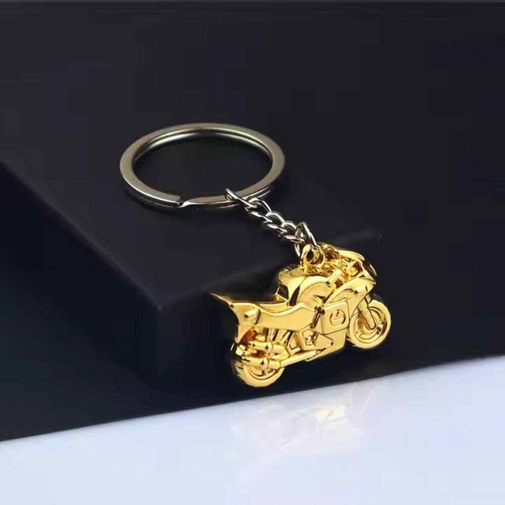 Złoty metalowy brelok w kształcie motocykla przymocowany do kółka na klucze, leży na ciemnym tle.