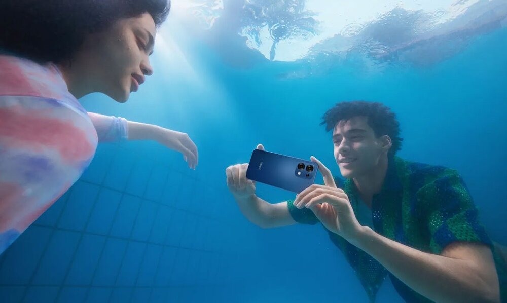 Dwie osoby znajdują się pod wodą, jedna z nich trzyma niebieski smartfon z widocznym logo OPPO. Tło stanowi basen z kafelkami.