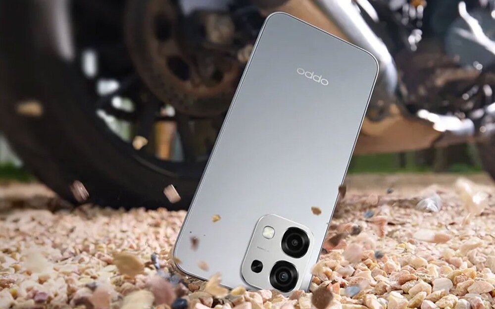 Smartfon w kolorze niebieskim leży na żwirowej powierzchni, w tle widoczne jest koło pojazdu. Na obudowie telefonu znajduje się logo \'oppo\' oraz moduł z trzema aparatami.