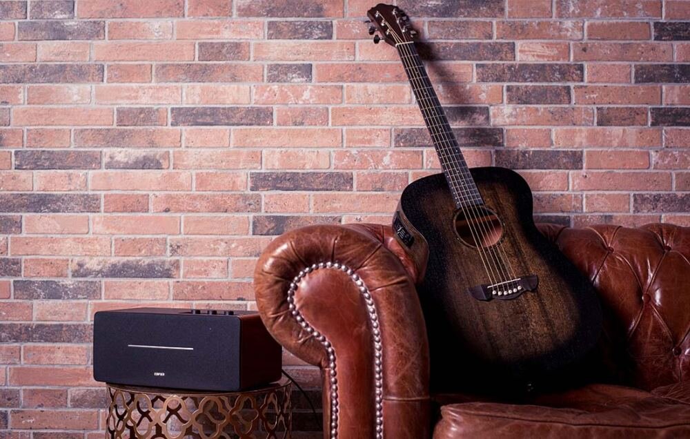 Na tle ceglanej ściany znajduje się brązowa, skórzana kanapa, na której oparta jest gitara akustyczna. Obok kanapy stoi stolik z czarnym głośnikiem, na którym widnieje logo \'EDIFIER\'.