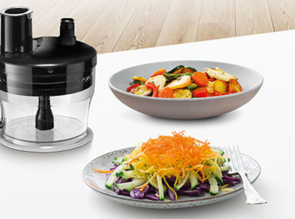 Na obrazie widoczny jest blender z przezroczystą misą i czarną pokrywą. Obok znajdują się dwa talerze z pokrojonymi warzywami.