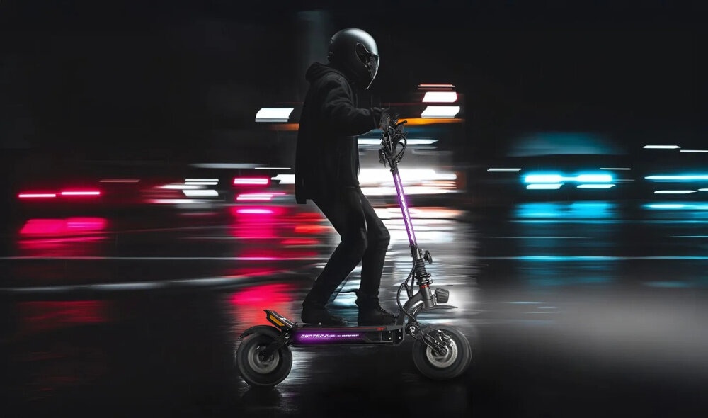 Osoba w kasku jedzie na czarnej hulajnodze elektrycznej po mokrej nawierzchni, w tle rozmyte światła miasta. Na hulajnodze widoczny jest napis \'RUPTOR R9\'.