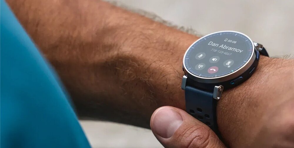 Osoba nosi smartwatch z okrągłym wyświetlaczem, na którym widoczny jest ekran połączenia z tekstem \'Dan Abramov\'. Pasek zegarka jest ciemny z perforacjami.