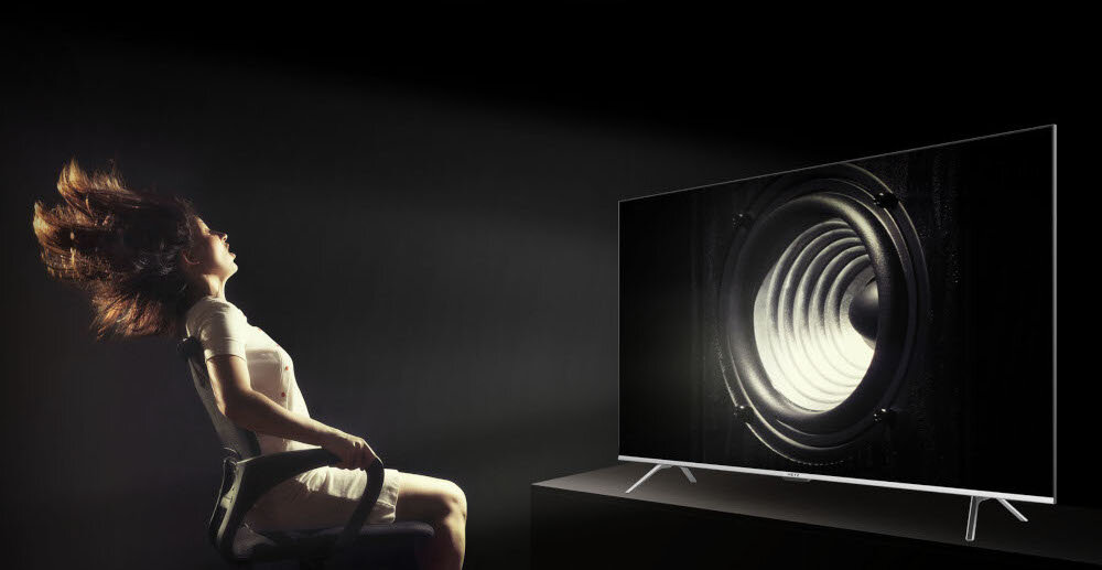 Osoba siedzi na krześle przed dużym telewizorem, na którym wyświetlany jest obraz głośnika. Włosy osoby są uniesione, jakby pod wpływem dźwięku.
