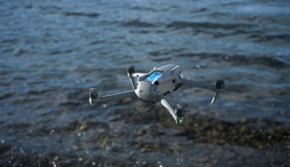 Dron unosi się nad wodą, widoczny jest jego szary korpus i cztery śmigła. W tle widać powierzchnię wody z lekkimi falami.