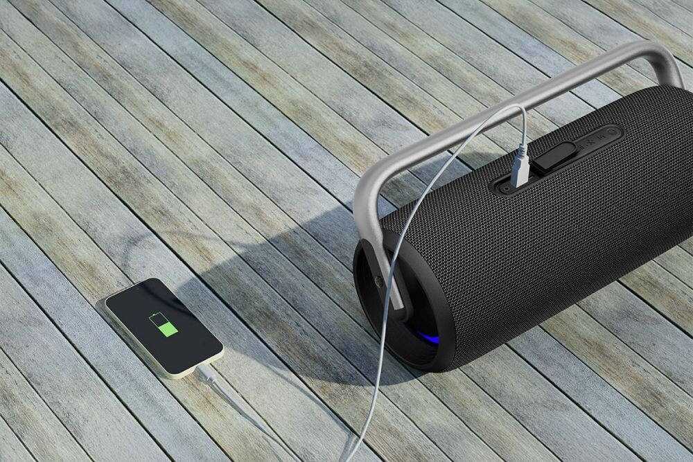 Na drewnianej podłodze znajduje się czarny głośnik mobilny z metalowym uchwytem, do którego podłączony jest kabel. Obok leży smartfon z wyświetlonym symbolem ładowania baterii.