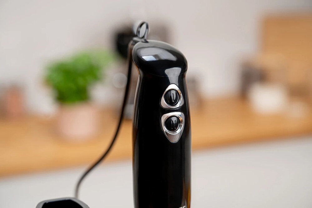 Czarny blender ręczny z dwoma przyciskami na uchwycie. W tle rozmyte elementy kuchenne, w tym zielona roślina w doniczce.