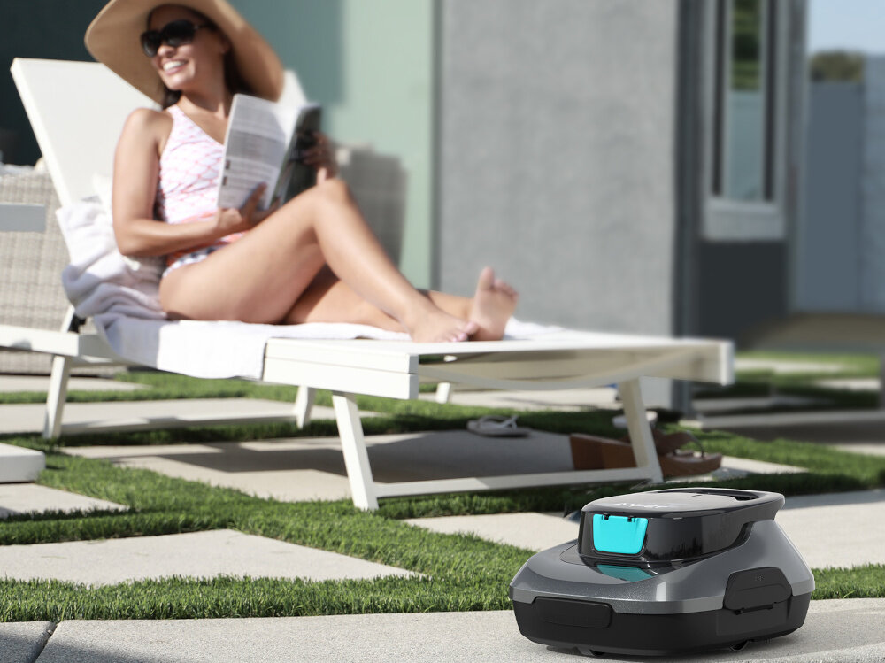 Na obrazie widać robota do czyszczenia basenów na tle osoby leżącej na leżaku. Robot ma nowoczesny, szary design z niebieskim elementem na górze.