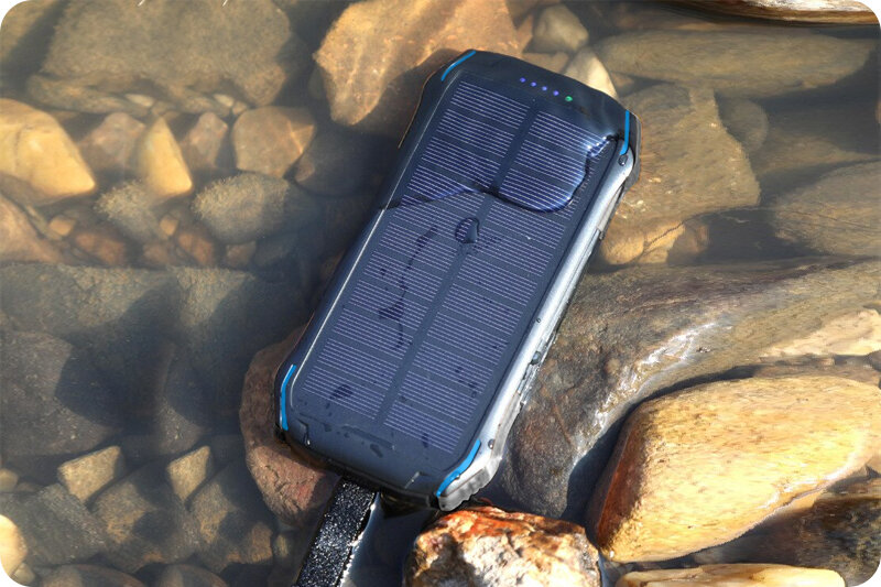 Na obrazie widać wodoodporny powerbank solarny zanurzony w wodzie, otoczony kamieniami. Urządzenie ma panel słoneczny na wierzchu i jest w kolorze czarnym z niebieskimi akcentami.