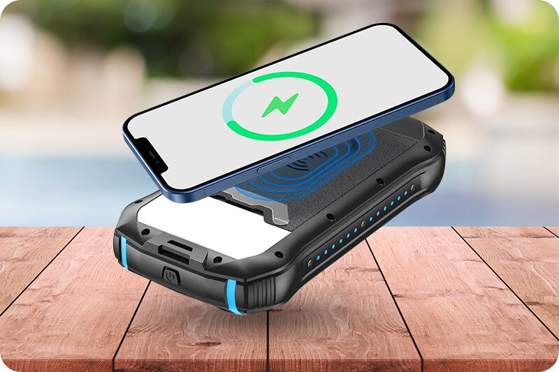 Na drewnianym stole leży czarny powerbank z niebieskimi akcentami, na którym unosi się smartfon z ikoną ładowania. W tle widoczny jest rozmyty krajobraz.