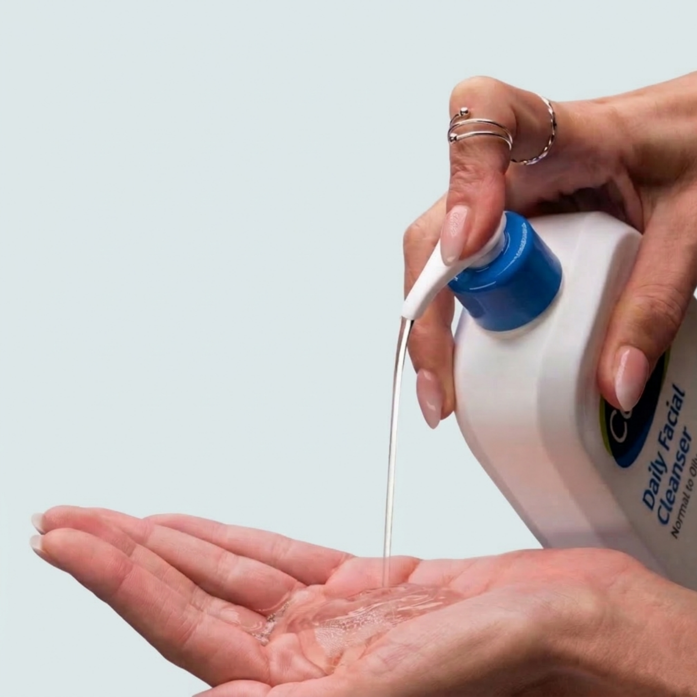 Osoba nalewa przezroczysty płyn z białej butelki z niebieskim korkiem na dłoń. Na butelce widoczny jest tekst: \'Daily Facial Cleanser Normal to Oily Skin\'.