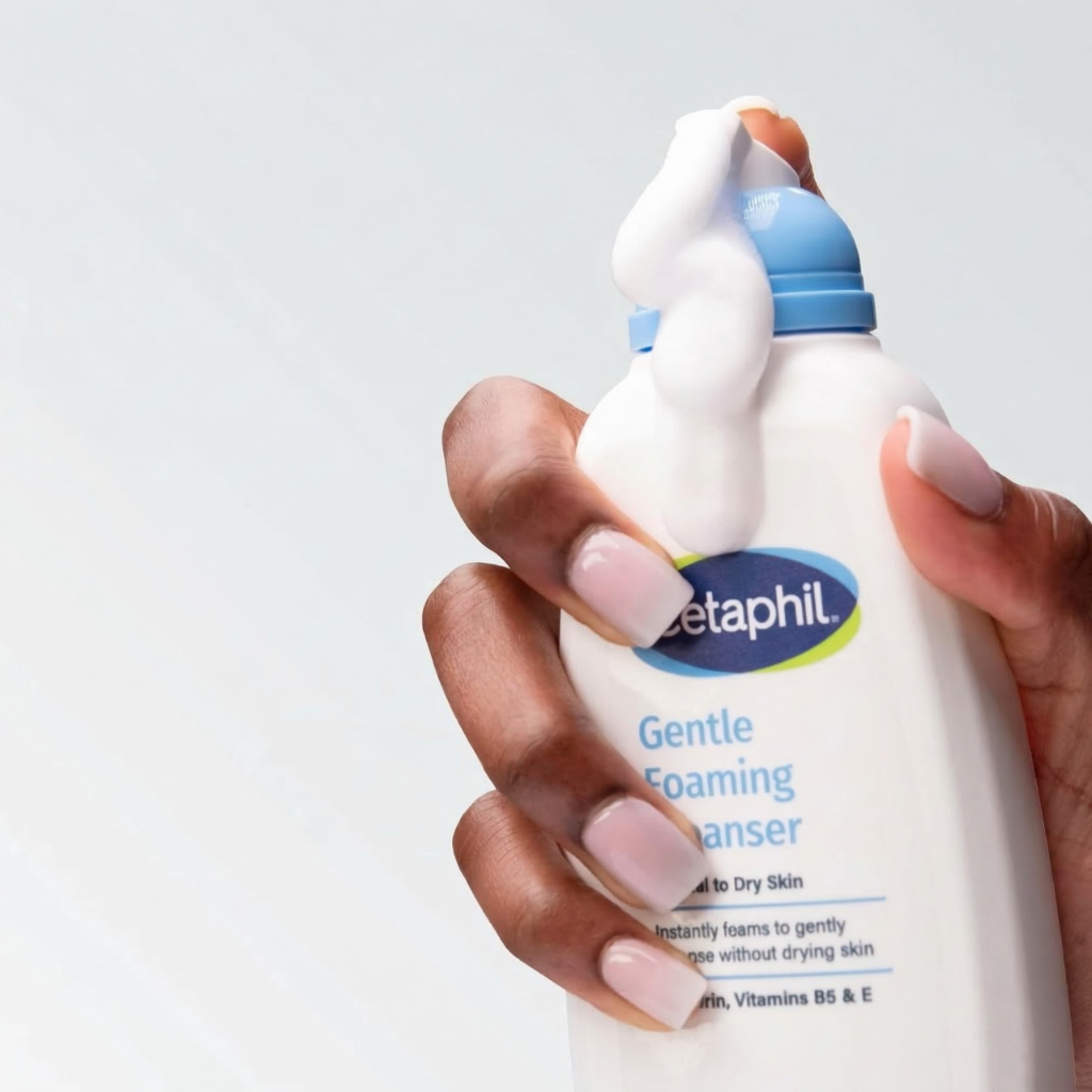 Osoba trzyma butelkę z pianką do mycia twarzy marki Cetaphil. Na etykiecie widnieje napis: \'Gentle Foaming Cleanser for Normal to Dry Skin Instantly foams to gently cleanse without drying skin Glycerin, Vitamins B5 & E\'.