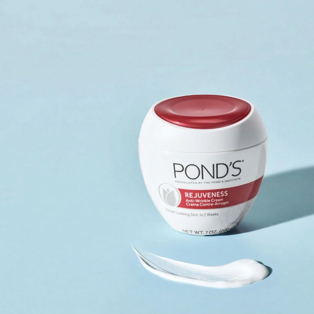 Na jasnoniebieskim tle znajduje się biały słoik z czerwonym wieczkiem, na którym widnieje napis \'POND\'S REJUVENESS Anti-Wrinkle Cream Crema Contre-Arrugas\'. Obok słoika znajduje się niewielka ilość białego kremu.