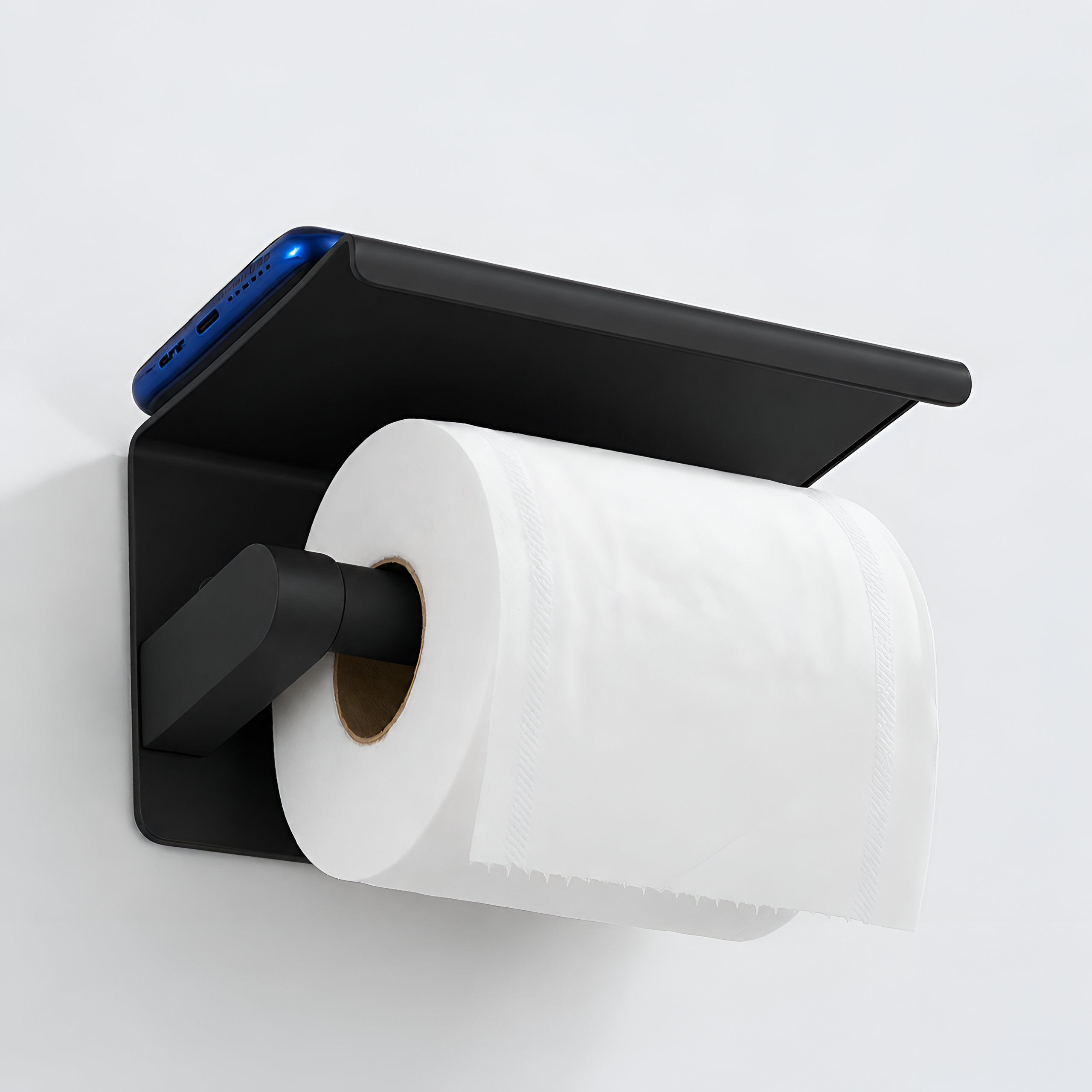 Czarny matowy uchwyt na papier toaletowy z półką, na której leży niebieski telefon. Na uchwycie znajduje się rolka białego papieru toaletowego.