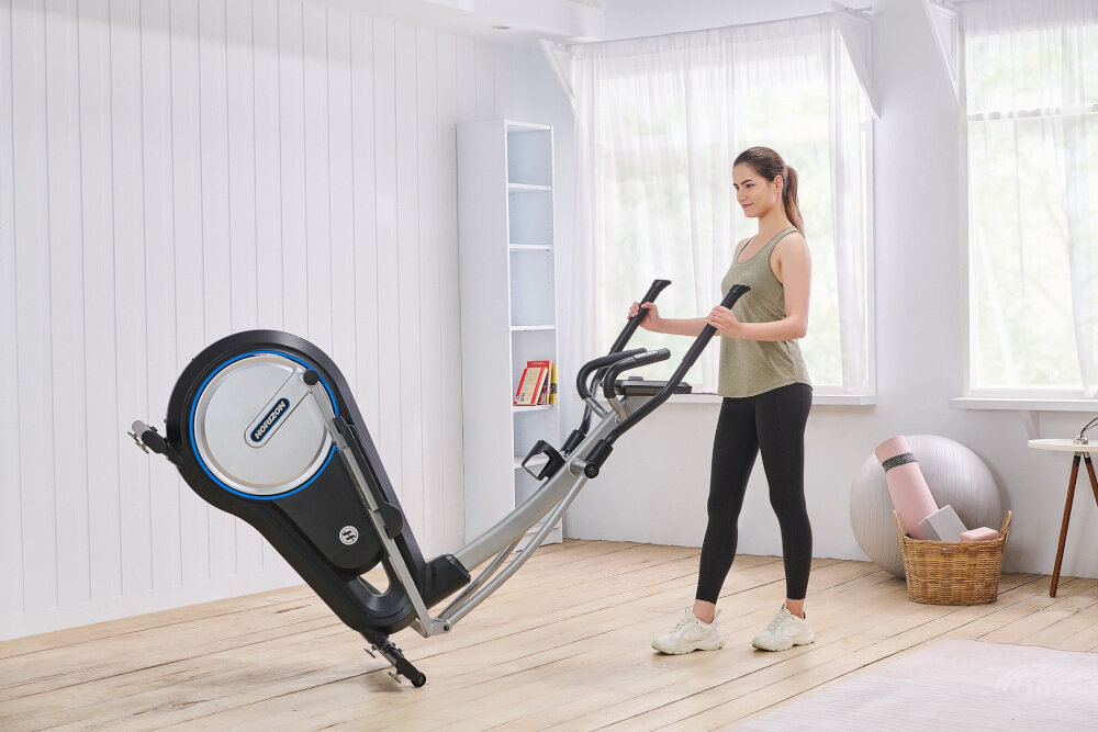 Kobieta stoi obok orbitreka eliptycznego w jasnym pomieszczeniu. Na urządzeniu widoczne jest logo \'HORIZON FITNESS\'.