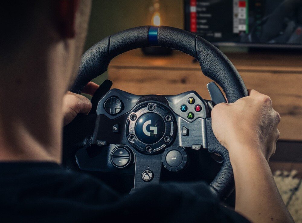 Osoba trzyma kierownicę do gier z widocznym logo \'G\' na środku. Na kierownicy znajdują się przyciski oznaczone literami A, B, X, Y oraz symbolem Xbox.