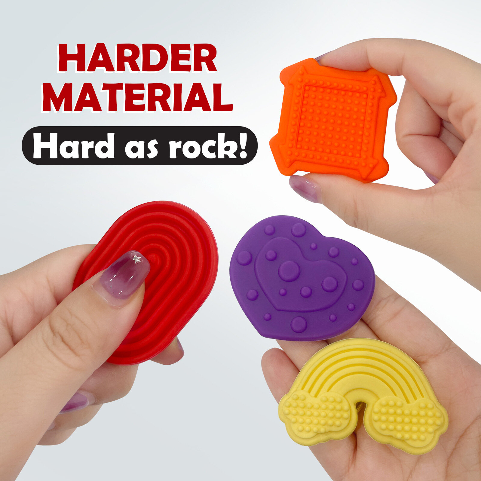 Na obrazie widoczne są cztery kolorowe, dotykowe kamyki sensoryczne o różnych kształtach i fakturach, trzymane w dłoniach. Tekst na obrazie: \'HARDER MATERIAL Hard as rock!\'.