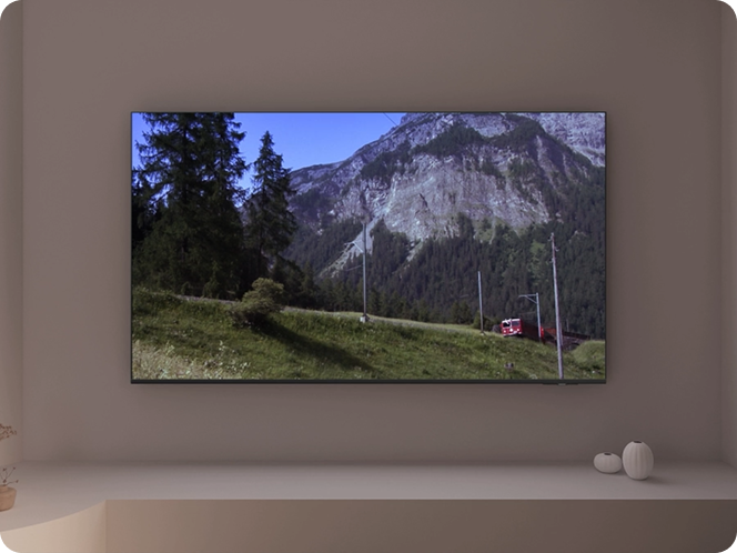 Telewizor wyświetla obraz górskiego krajobrazu z drzewami i pociągiem na torach. W tle widać góry i niebo.