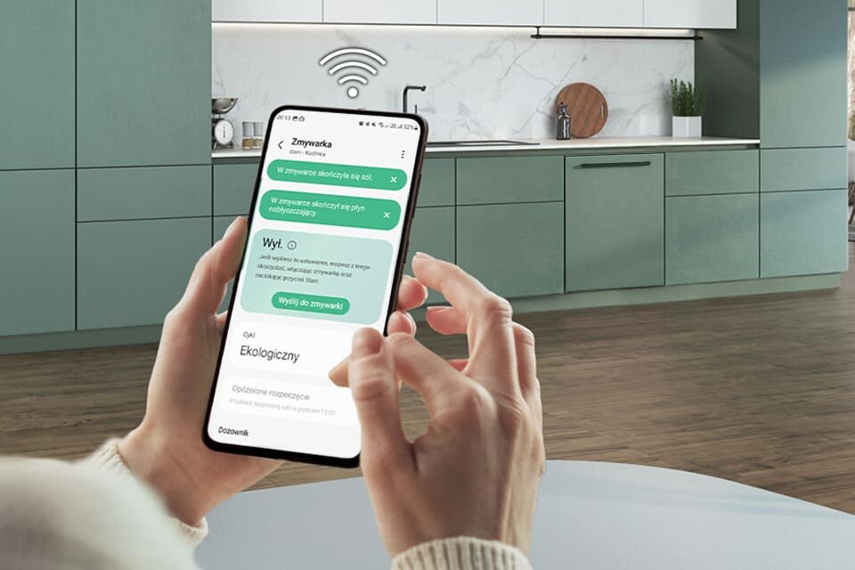 Osoba trzyma smartfon z aplikacją do zdalnego sterowania zmywarką, na ekranie widoczne są opcje: \'W zmywarce skończyły się sól i nabłyszczacz\', \'Wył. O\', \'Ekologiczny\'. W tle znajduje się nowoczesna kuchnia z zielonymi szafkami i marmurowym blatem.
