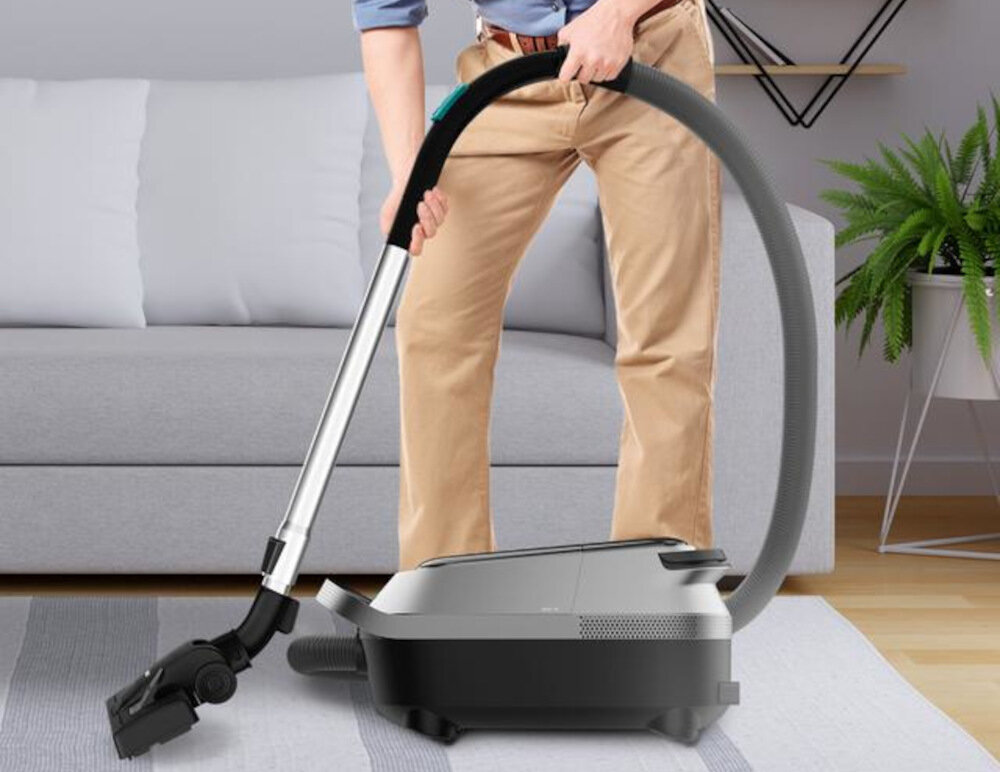 Osoba używa odkurzacza z metalową rurą i elastycznym wężem na tle szarej kanapy i rośliny w doniczce. Odkurzacz ma nowoczesny design i jest w kolorze czarnym z szarymi elementami.