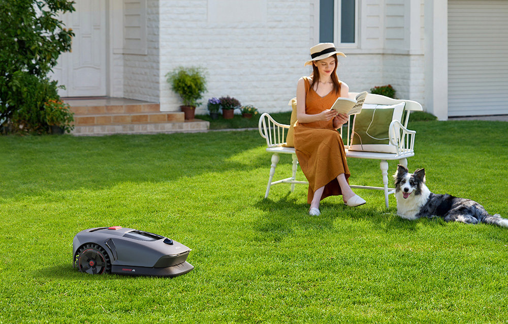 Na trawniku znajduje się robot koszący, a w tle kobieta siedzi na ławce z książką obok psa. W tle widoczny jest dom z białą fasadą i rośliny w doniczkach.