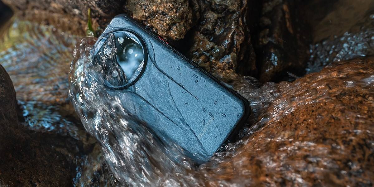 Smartfon w kolorze złotym leży w strumieniu wody, otoczony kamieniami. Na obudowie widoczne są krople wody.