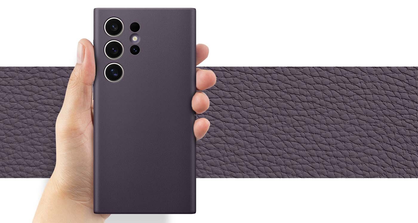 Vegan Leather Case chroni Twój telefon przed uszkodzeniami Osoba trzyma ciemnoszare etui na telefon z trzema dużymi i jednym małym otworem na aparaty. W tle widoczna jest tekstura przypominająca skórę w podobnym kolorze.