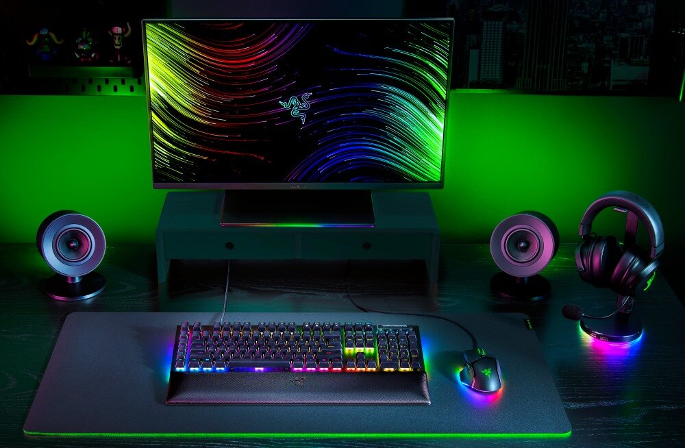 Na biurku znajduje się klawiatura z podświetleniem RGB, myszka oraz monitor z kolorową grafiką i logo Razer. Obok monitora stoją dwa głośniki i zestaw słuchawkowy na stojaku, również z podświetleniem RGB.