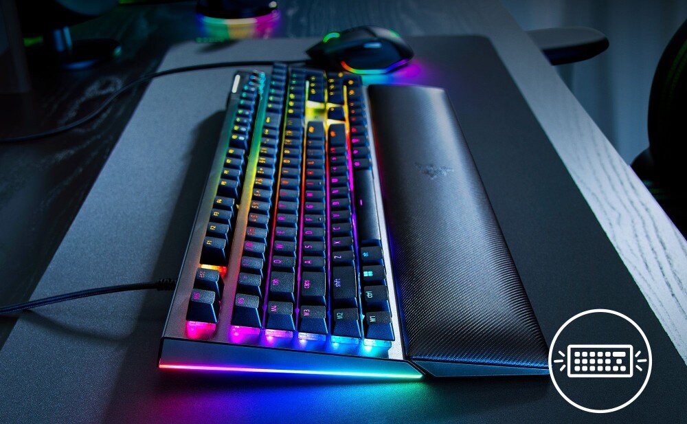 Na obrazie widoczna jest klawiatura z podświetleniem RGB oraz mysz komputerowa na ciemnym biurku. W prawym dolnym rogu znajduje się ikona klawiatury.